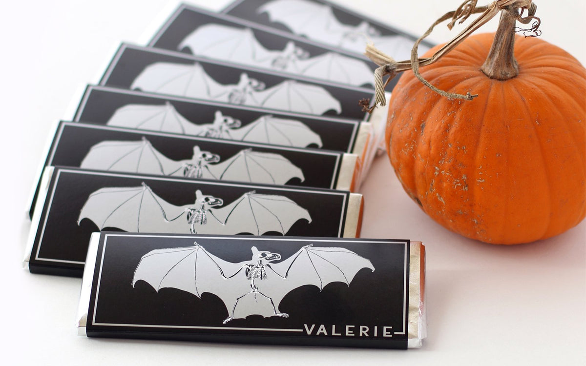 A stack of Bat Bars in black and silver wrappers, arranged next to a rustic pumpkin for a festive seasonal display.